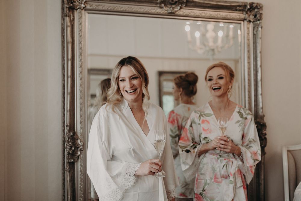 Two women in white robes sharing a joyful moment in front of an ornate mirror.