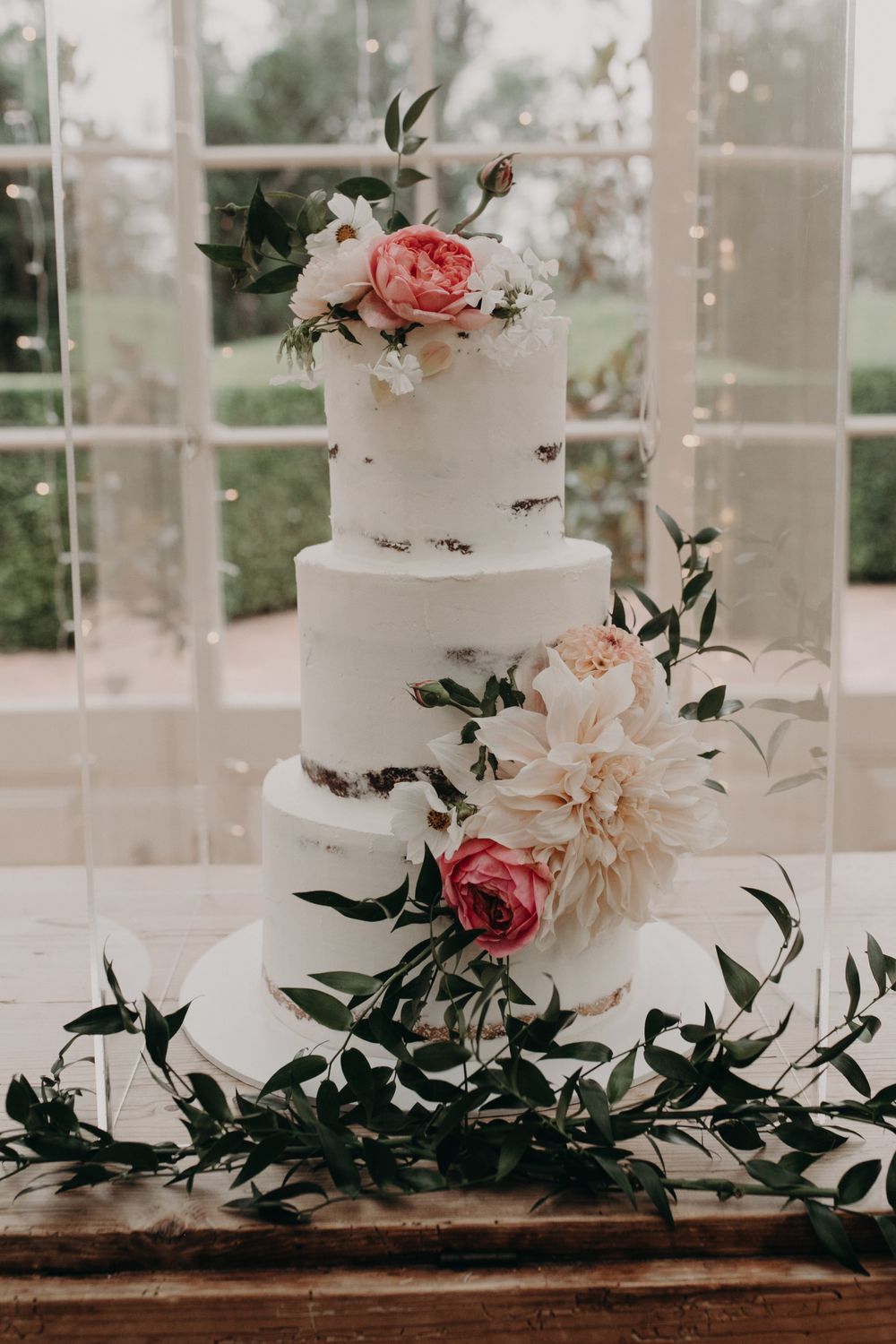 Three-tiered white wedding cake decorated with pink roses and greenery against a window backdrop.