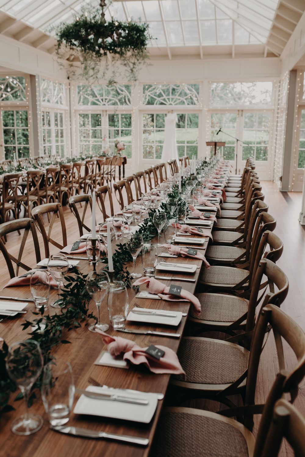 Elegant wedding reception table setting with greenery runners and cross-back chairs in a bright conservatory.