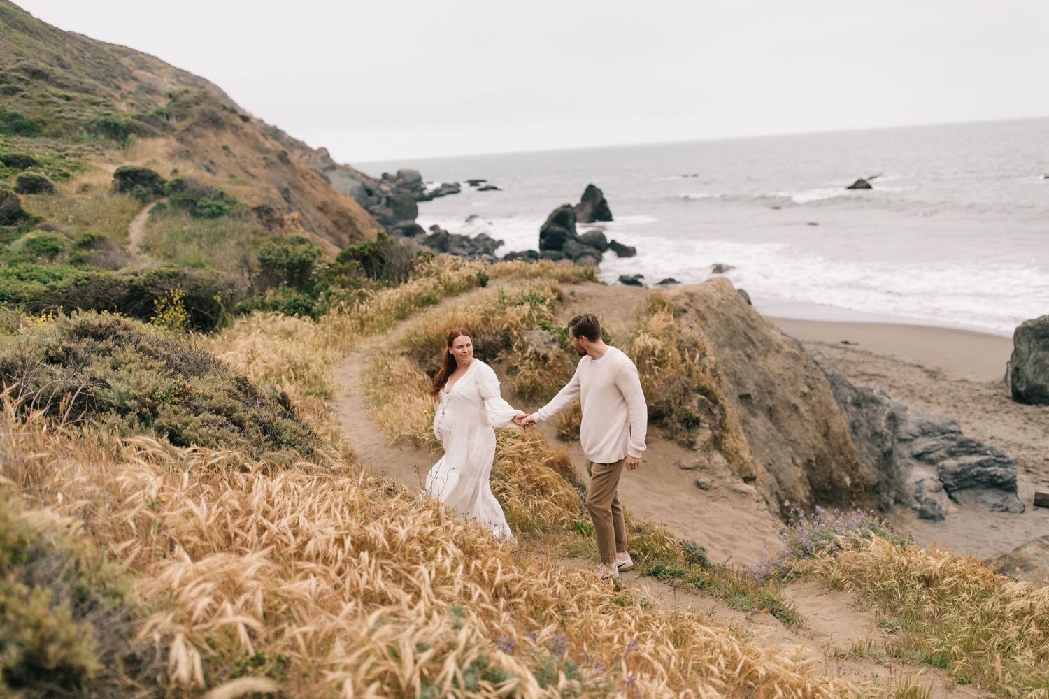 A couple walks along a scenic coastal cliff path wearing light colored clothing.