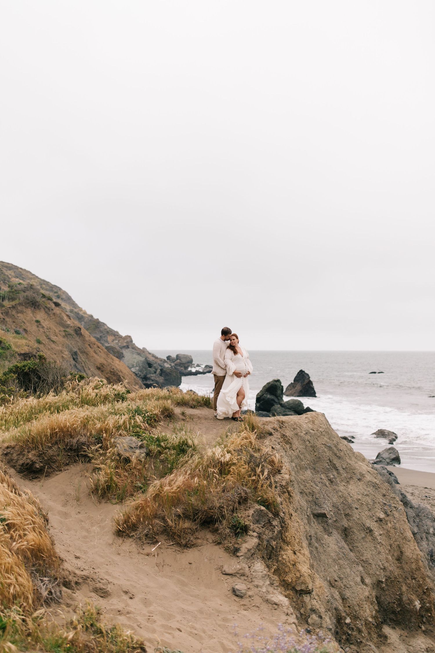 A romantic couple stands on a dramatic coastal cliff overlooking the ocean.