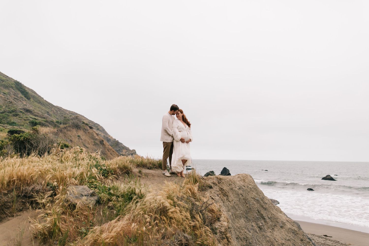 A couple embraces on a coastal cliff with ocean waves crashing below.
