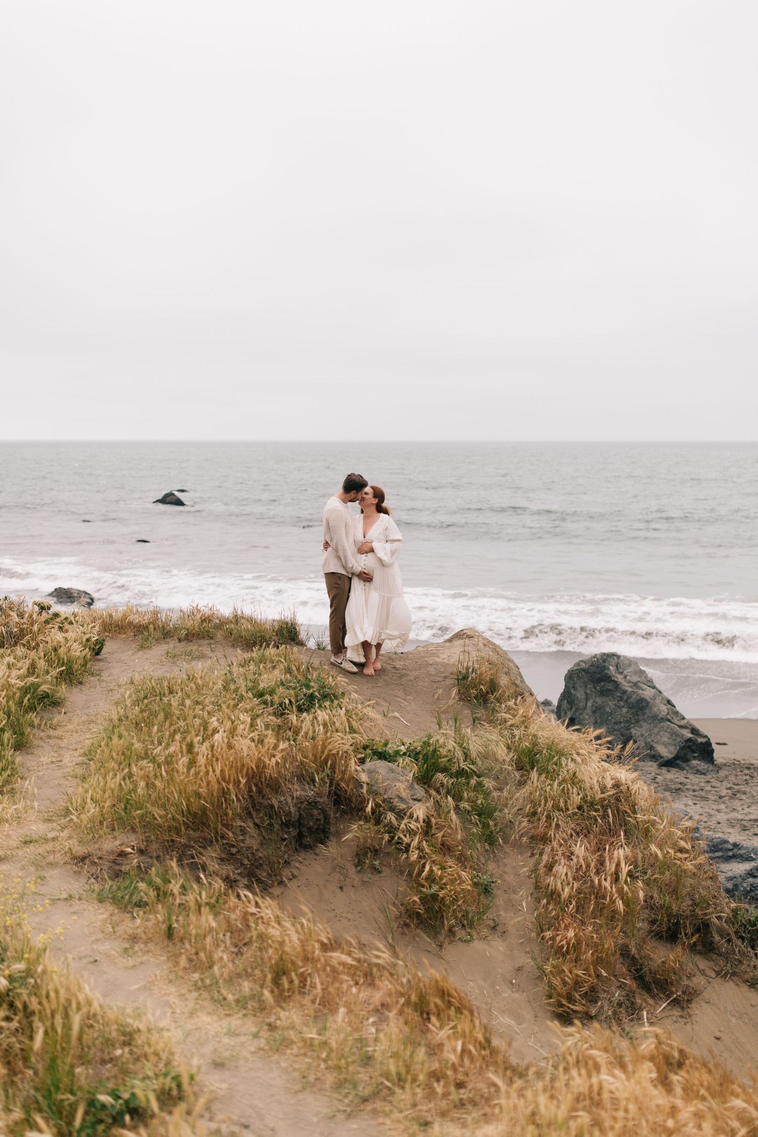 Couple standing on a grassy coastal bluff with ocean waves crashing below.