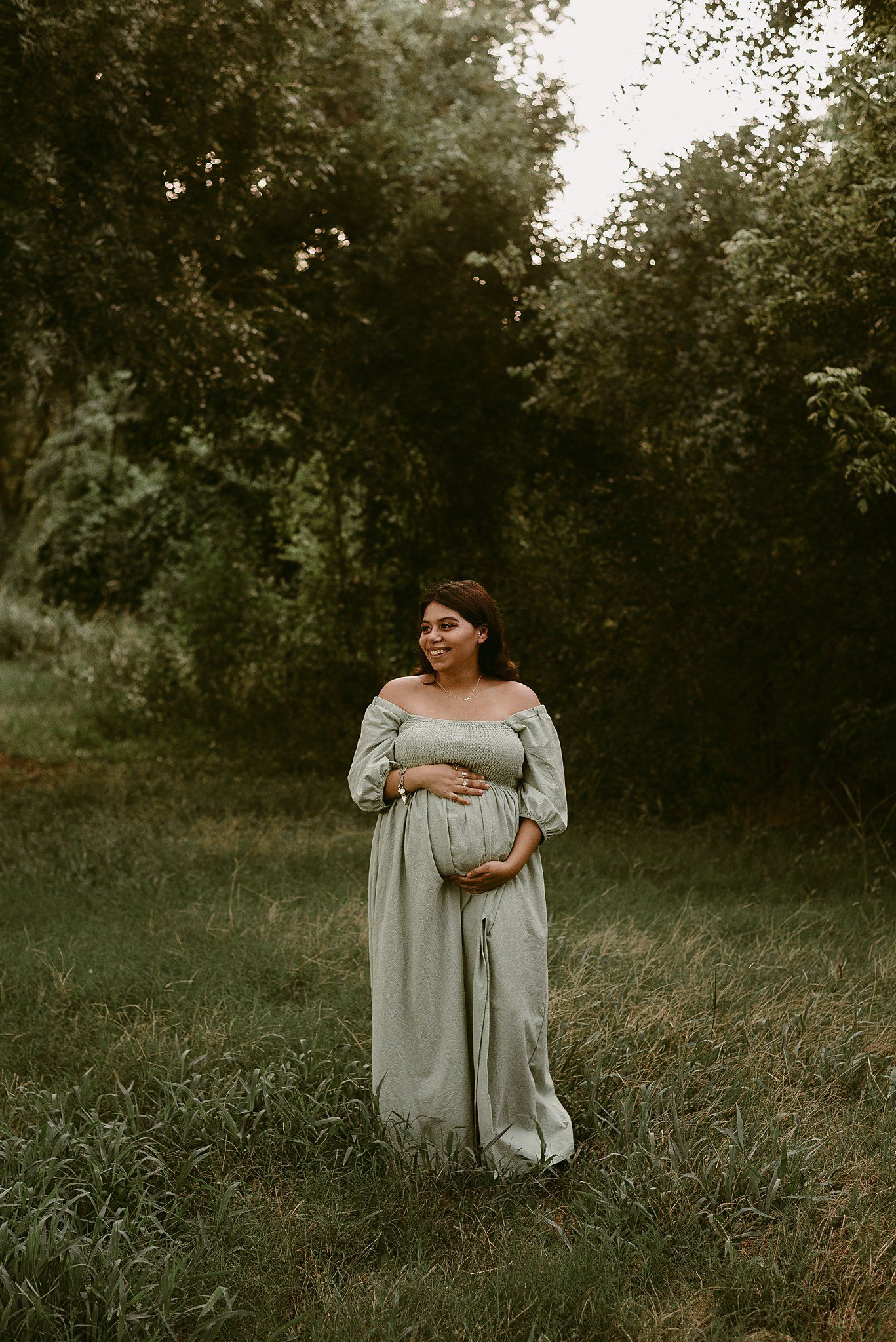 Expectant mother in off-shoulder grey dress poses in natural outdoor setting.