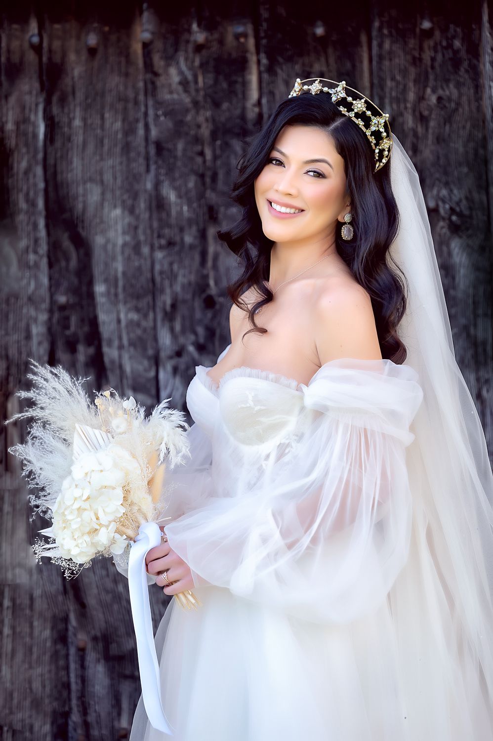Bride in off-shoulder white gown with crown holds dried flower bouquet against rustic backdrop.