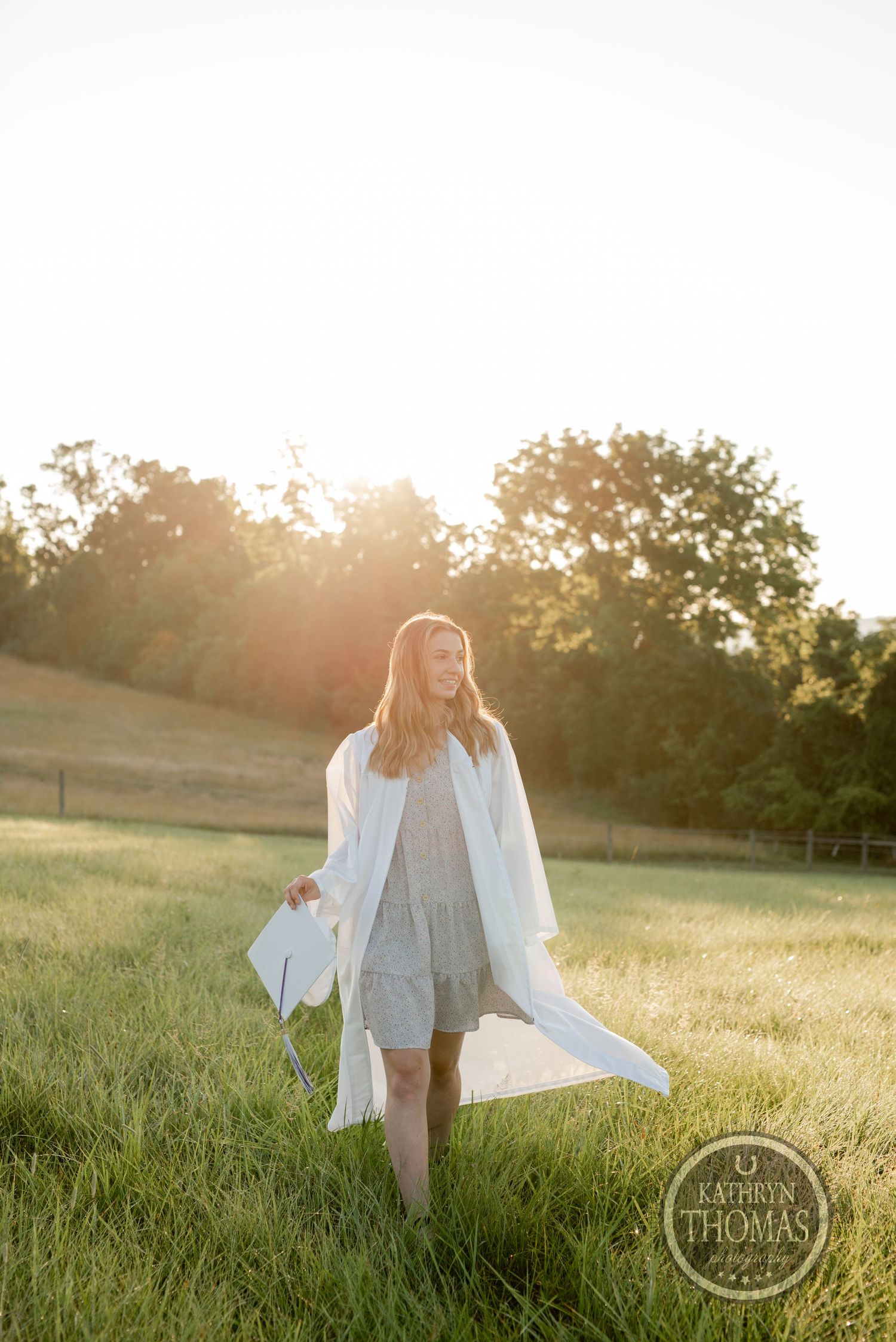 Graduate walking through sunlit field in flowing white graduation gown at sunset.