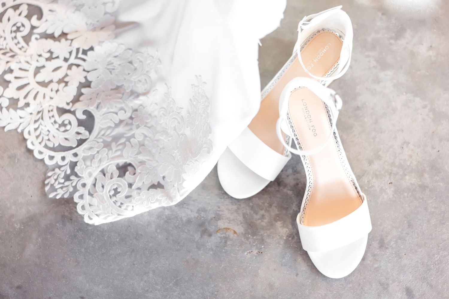Elegant white bridal sandals and delicate lace veil arranged on gray concrete surface.