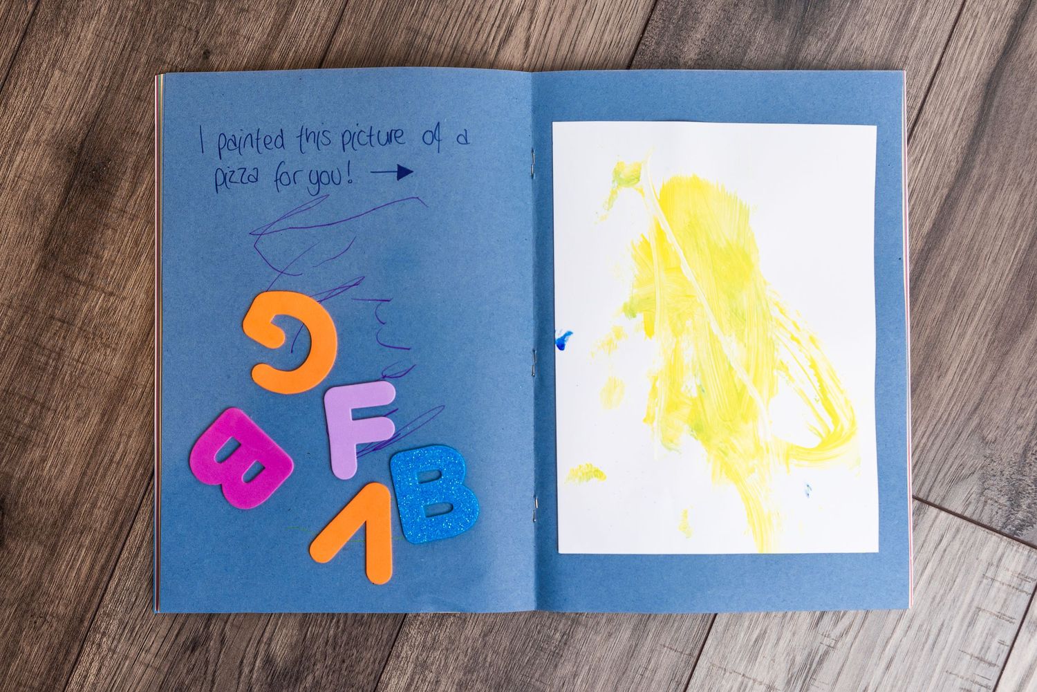 Open children's art book showing colorful letter cutouts and yellow drawing on blue paper.
