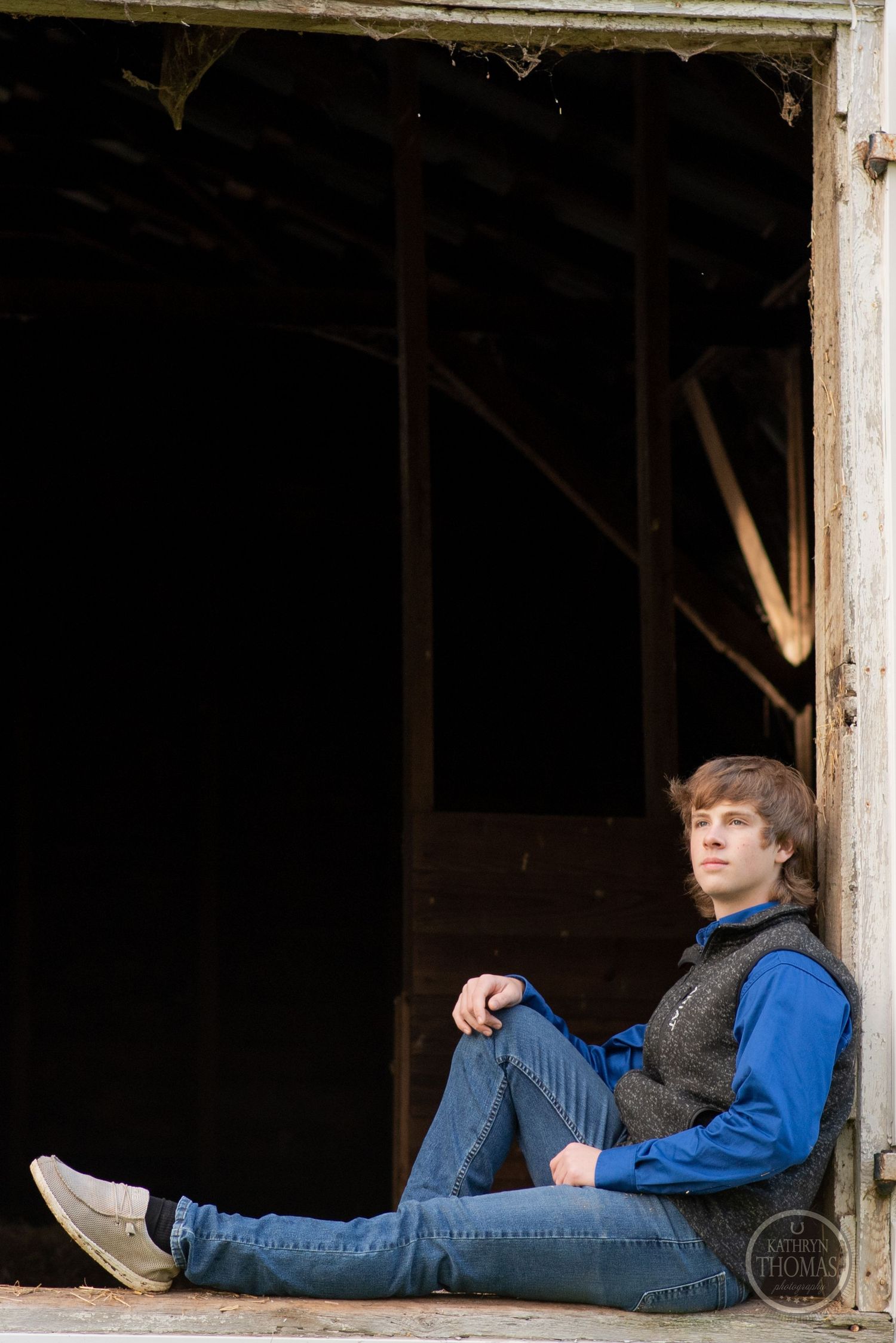 Person in blue jacket and jeans sits casually in rustic doorway of white barn.