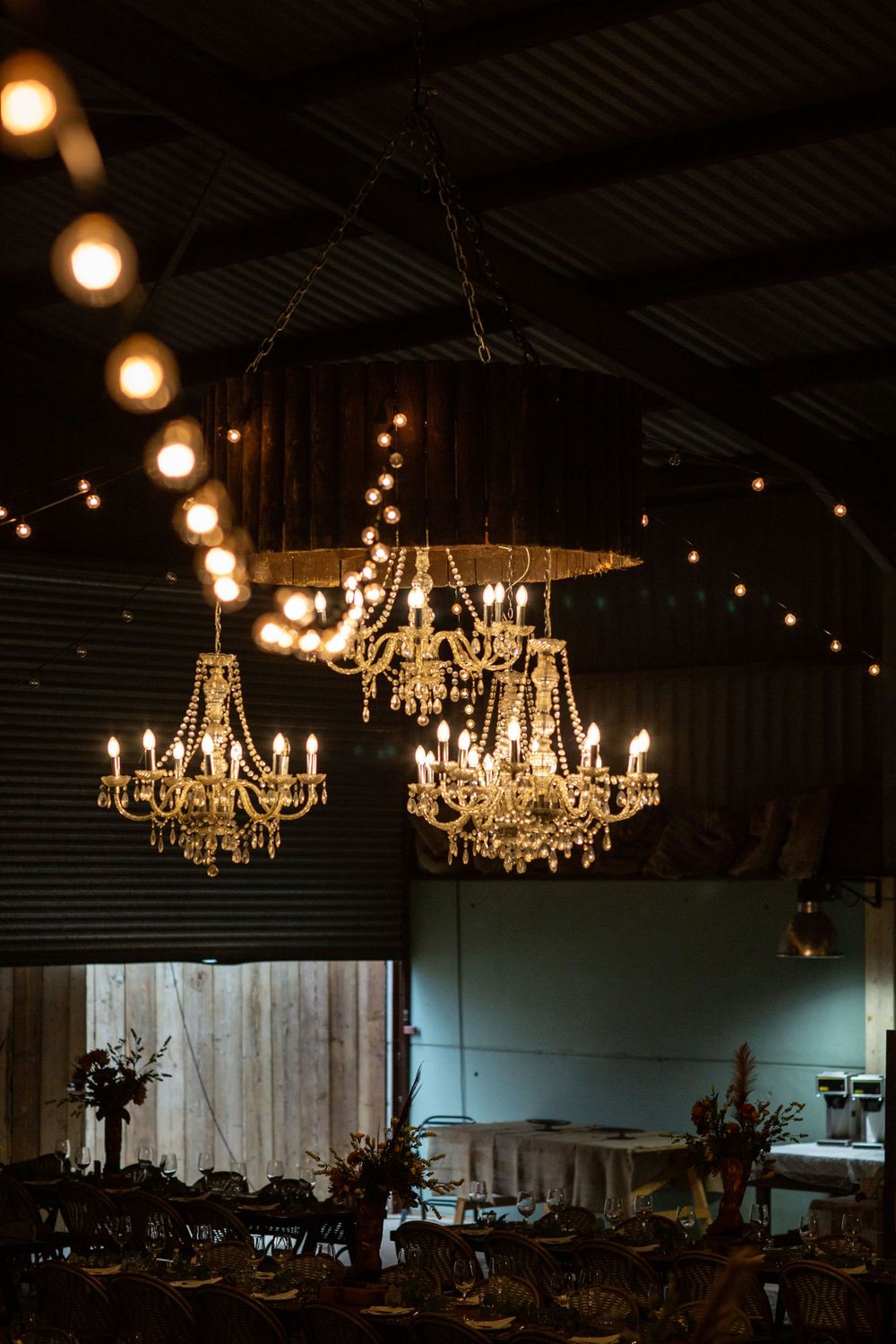 Three crystal chandeliers hanging in an industrial barn space with string lights.