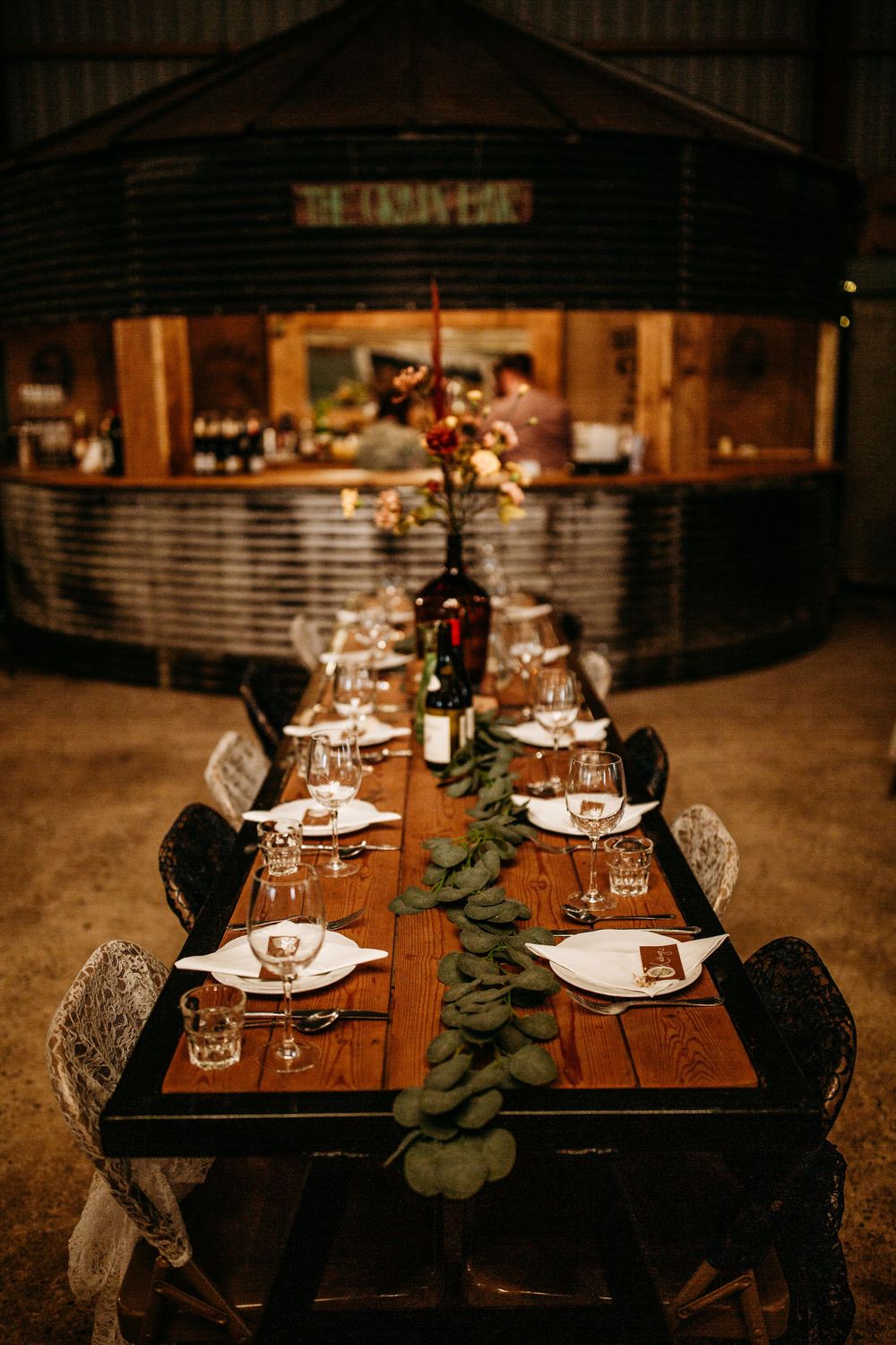 Elegant rustic dining table set with wine glasses and eucalyptus garland runner in a cozy barn setting.