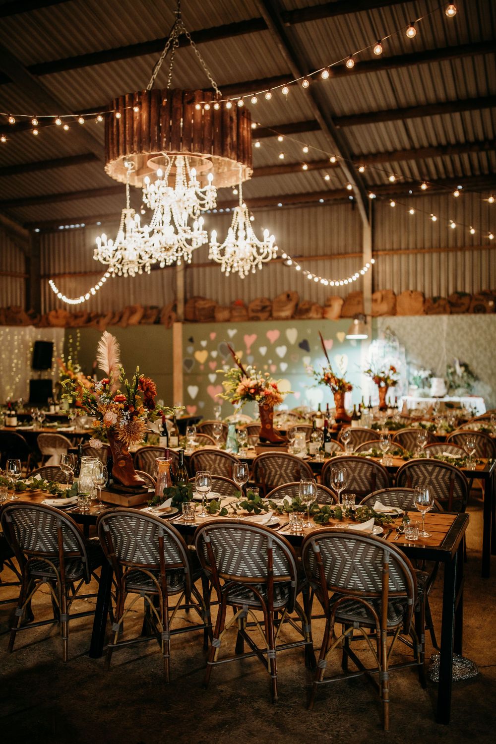 Romantic barn reception with crystal chandeliers and bistro lights over long wooden tables with French cafe chairs.