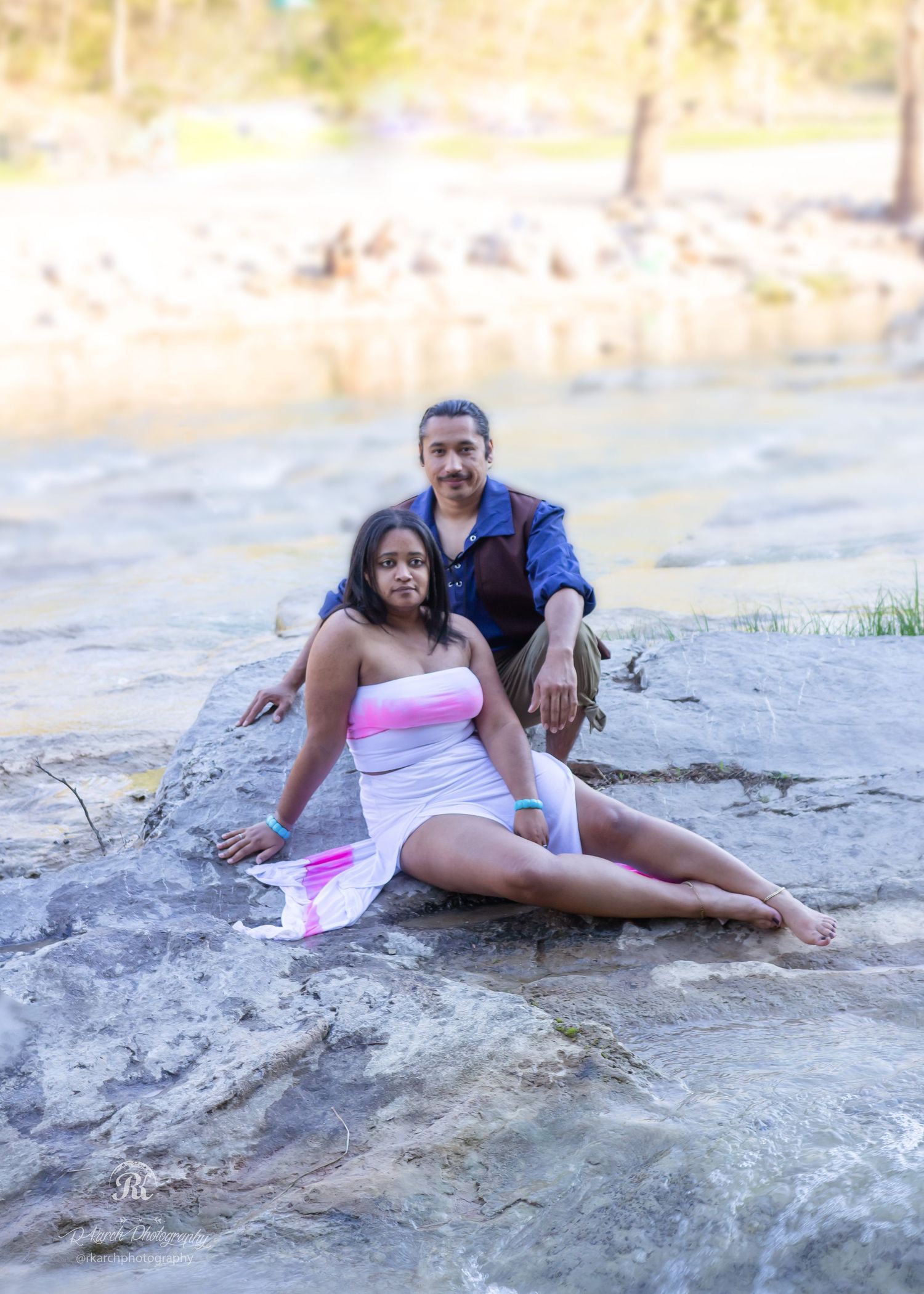 A couple relaxes together on rocky outcrop near flowing water on sunny day.
