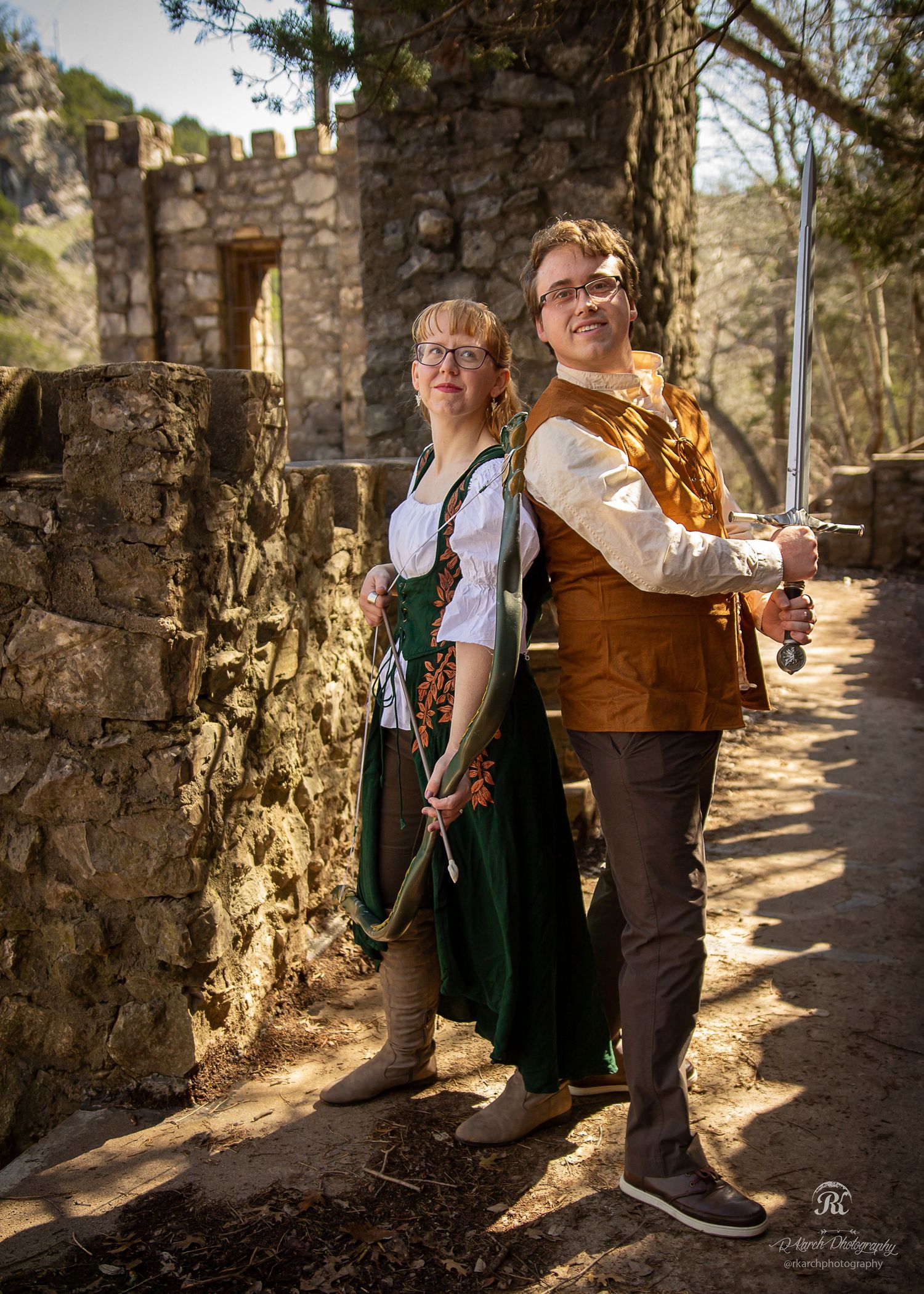 Two people in traditional Bavarian outfits pose back-to-back near stone castle ruins.