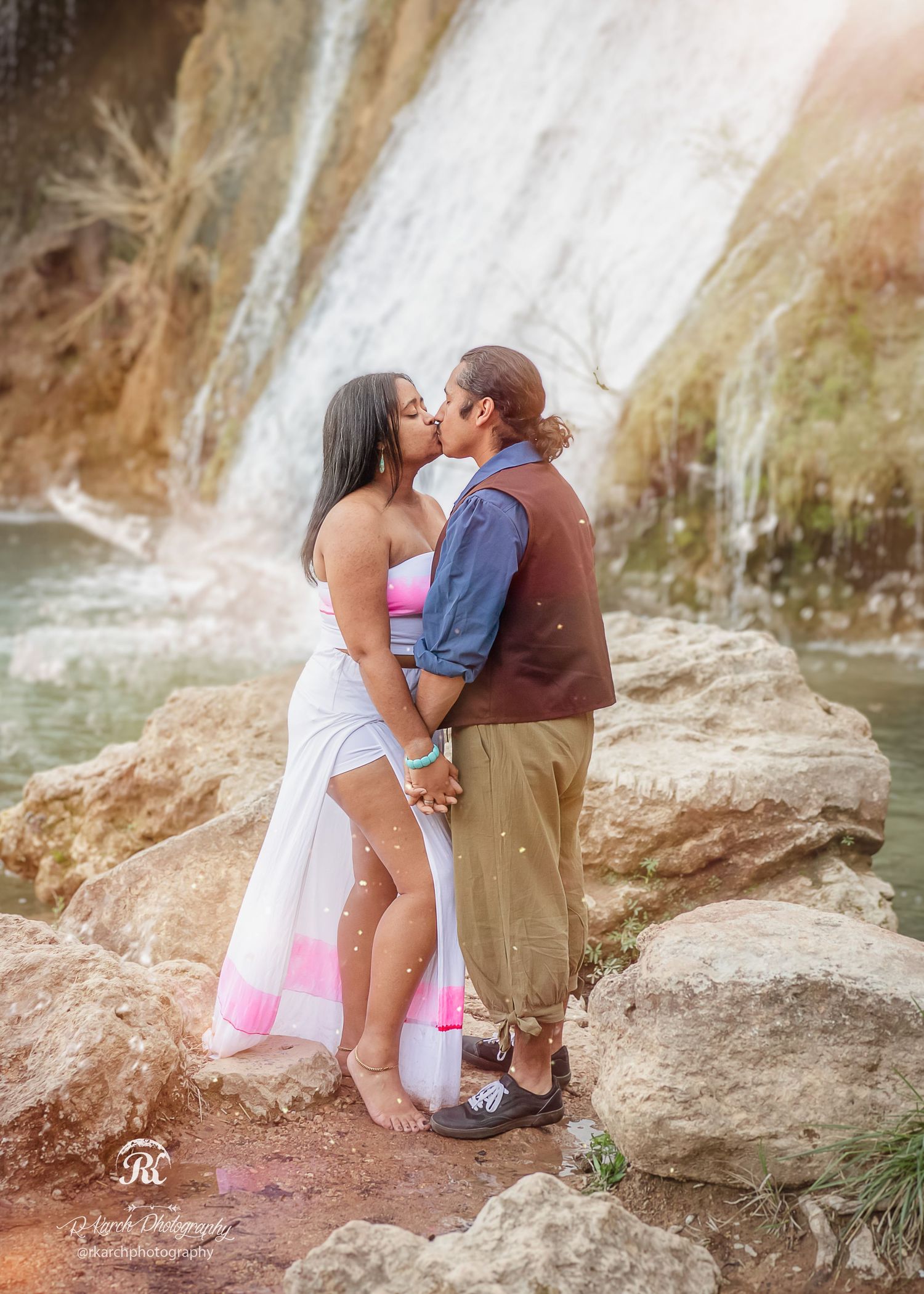 Romantic couple sharing a tender kiss in front of a cascading waterfall on rocky terrain.