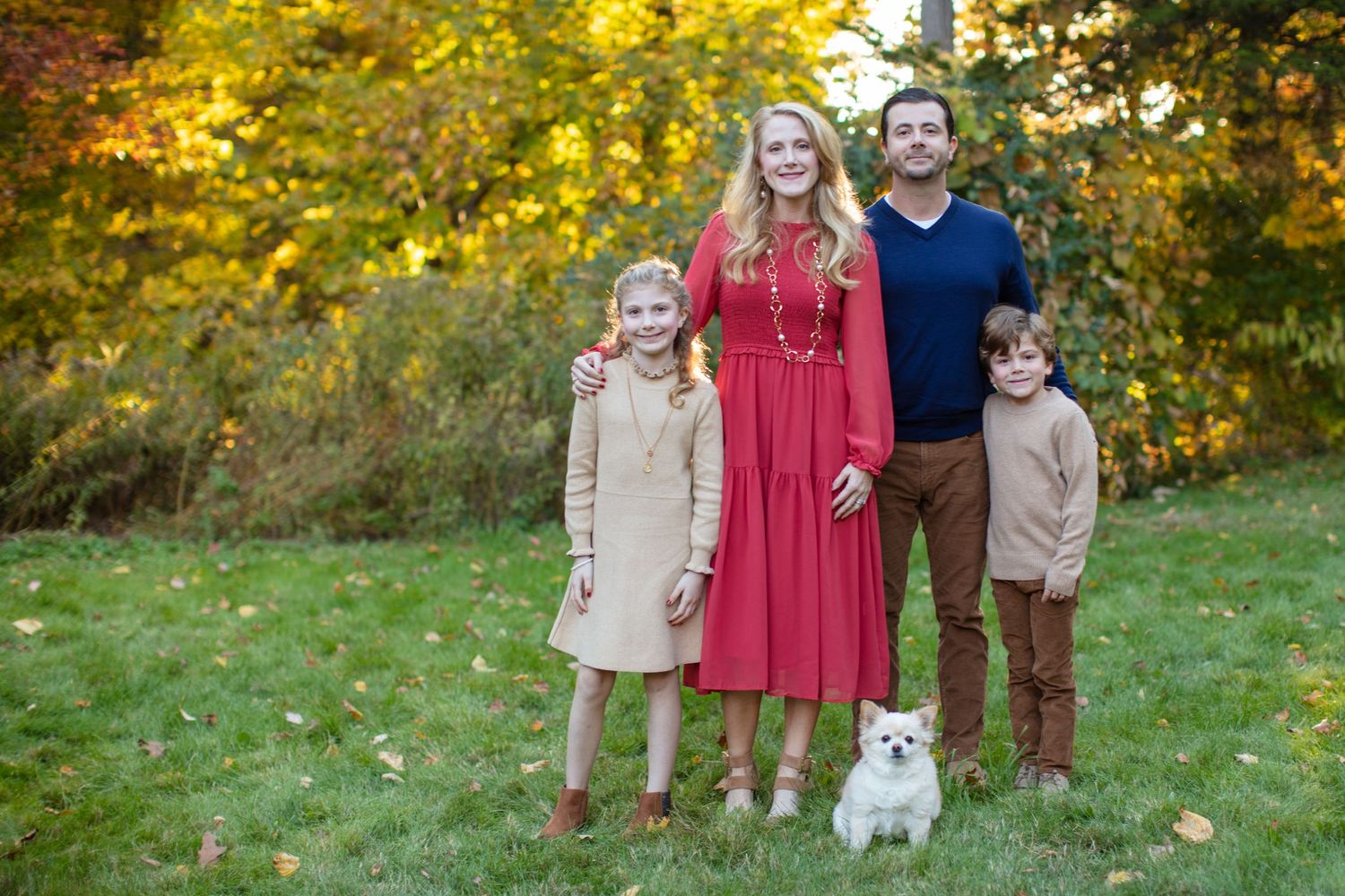 Family and small white dog pose together in autumn setting with golden trees behind.