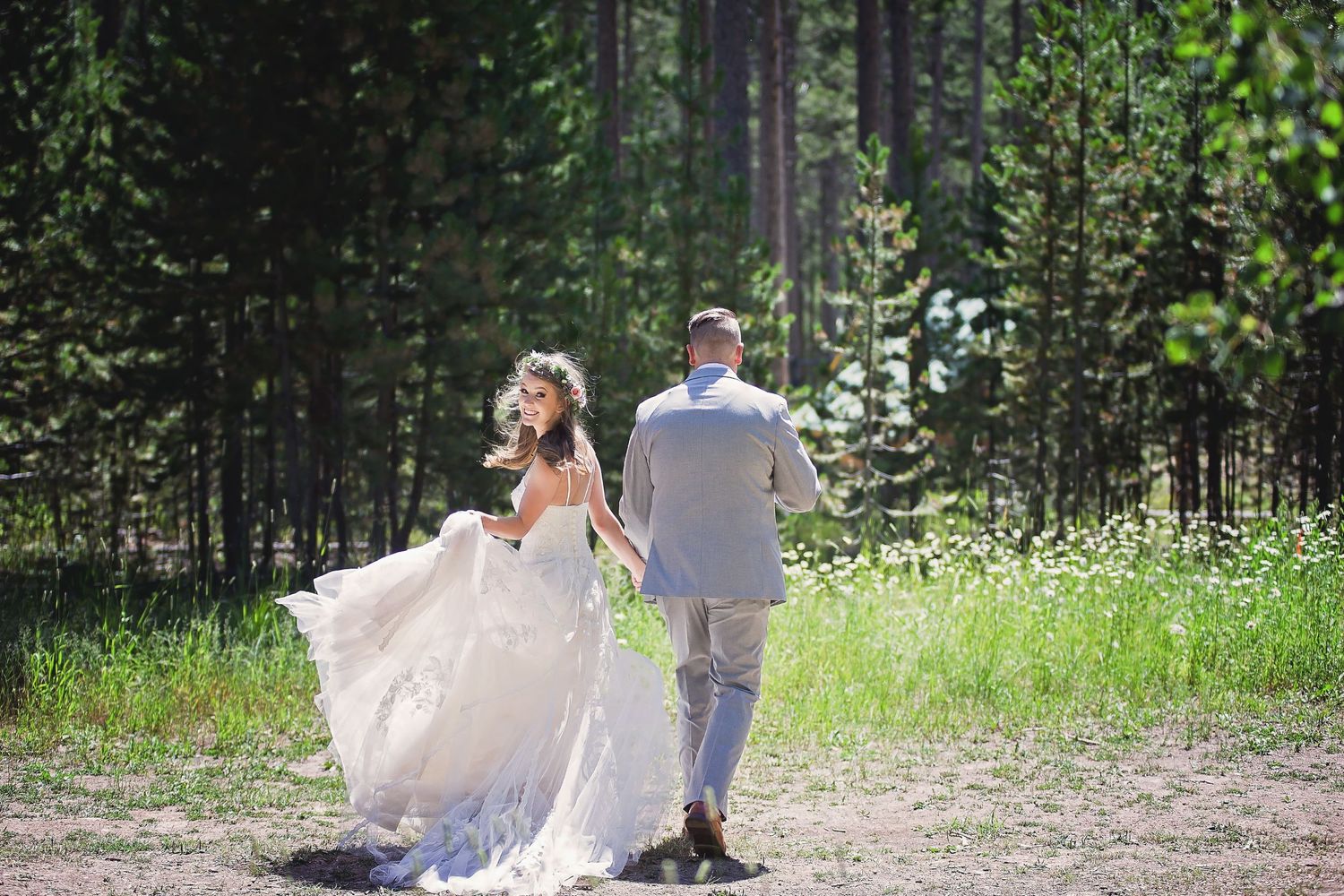 A couple in wedding attire walks through a serene forest setting with sunlit trees in the background.