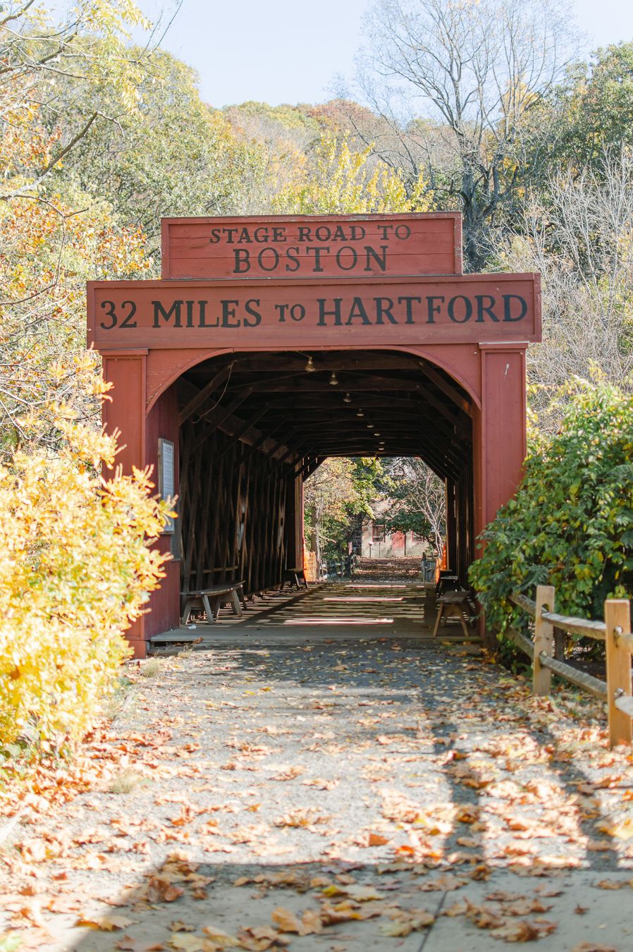 Historic red covered bridge entrance reading Stage Road to Boston, 32 Miles to Hartford in autumn.