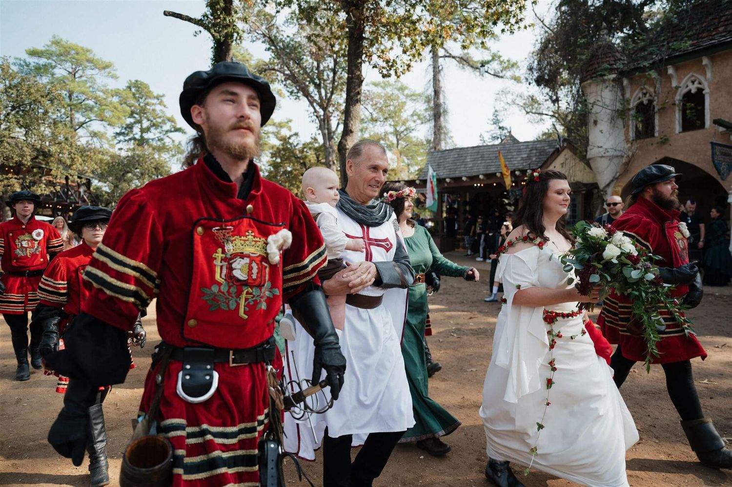 A Renaissance festival wedding procession with guards in red Tudor uniforms walking outdoors.