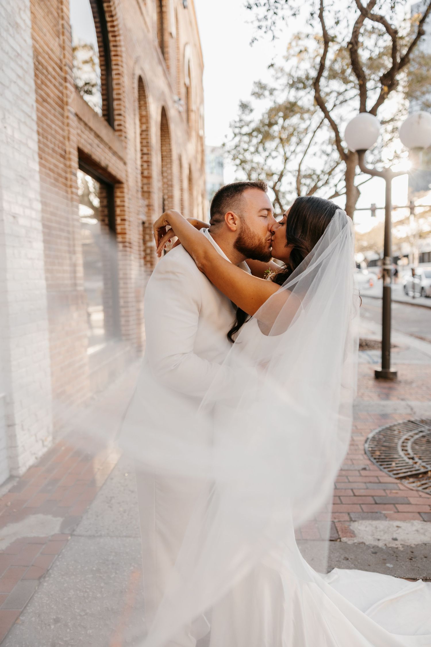 Bride and groom sharing a passionate kiss on a brick sidewalk, veil flowing in the breeze.