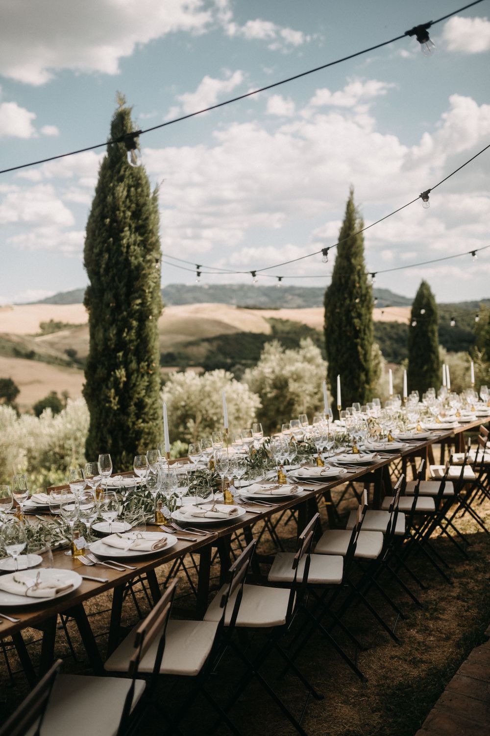 Long banquet tables set for wedding dinner under string lights with cypress trees and rolling hills.