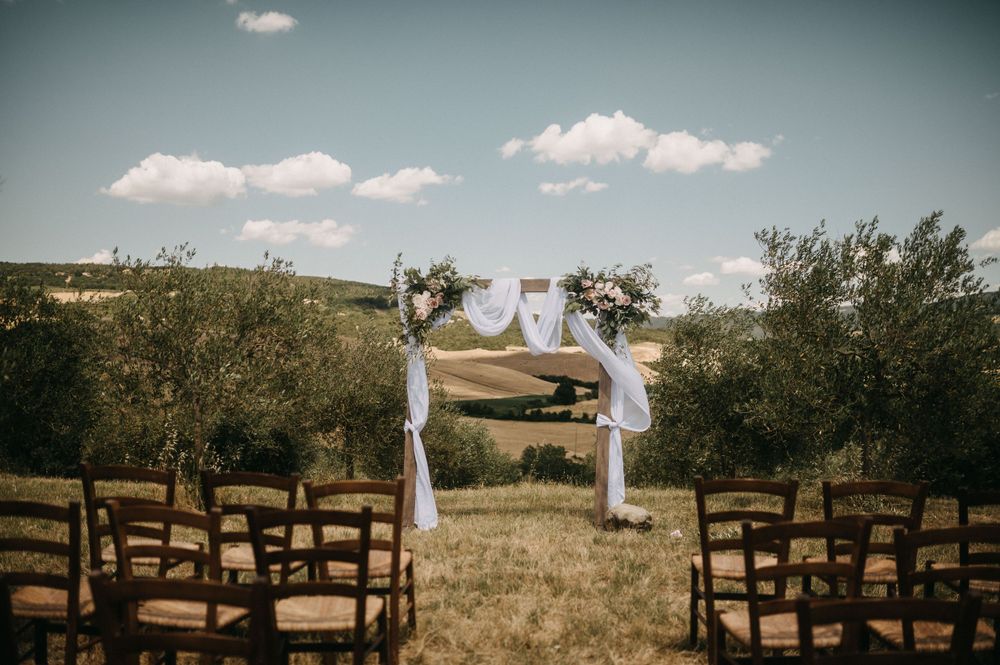 Rustic outdoor wedding ceremony setup with wooden benches and white fabric arch in a meadow.