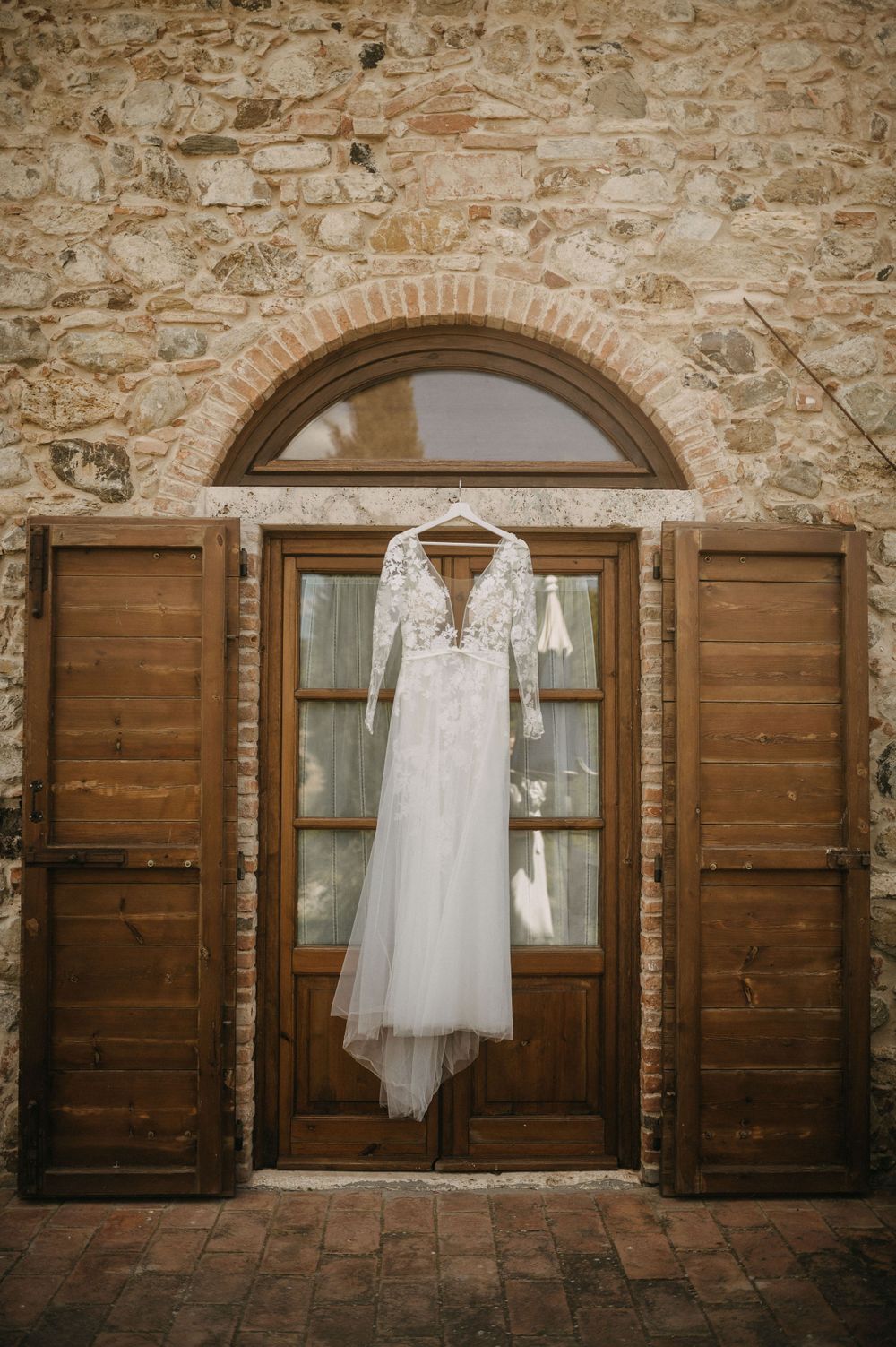 Elegant lace wedding dress hanging on wooden door with stone wall backdrop.