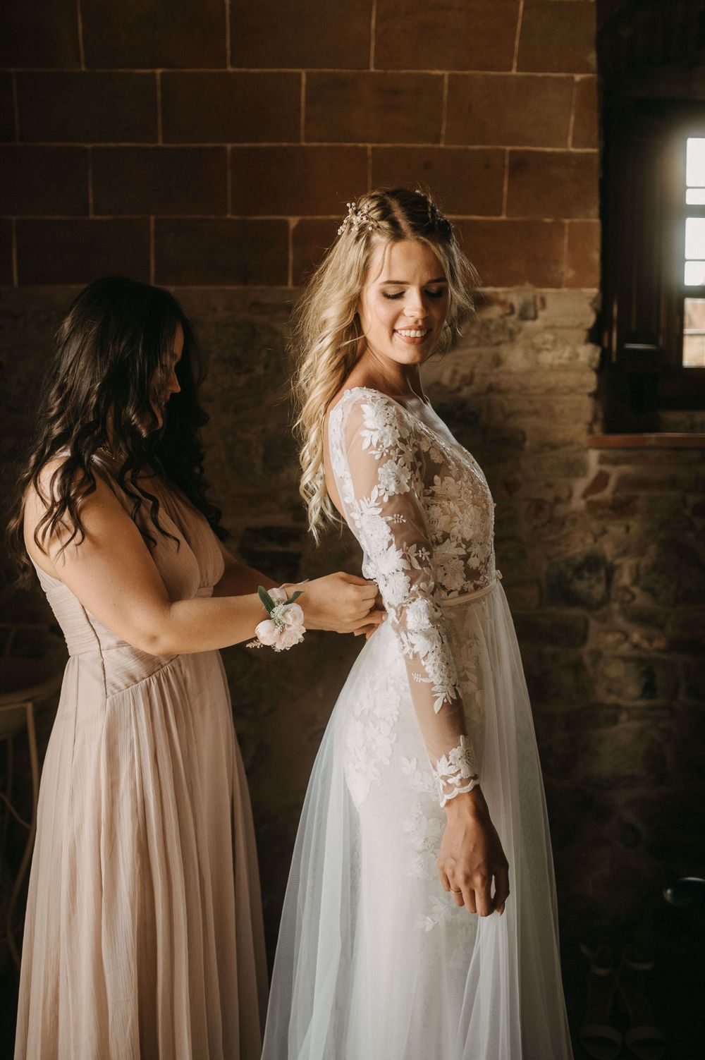 Bride in lace dress getting final adjustments from bridesmaid against brick wall.