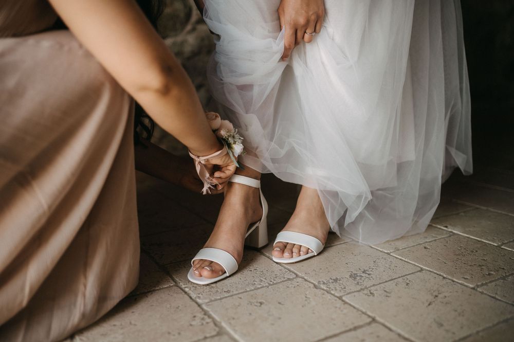 Close-up of hands fastening white bridal sandals on stone floor.