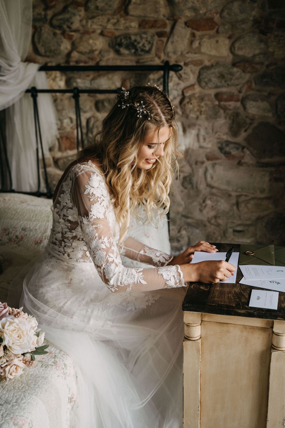 Bride in lace gown writing at vintage desk with roses nearby.