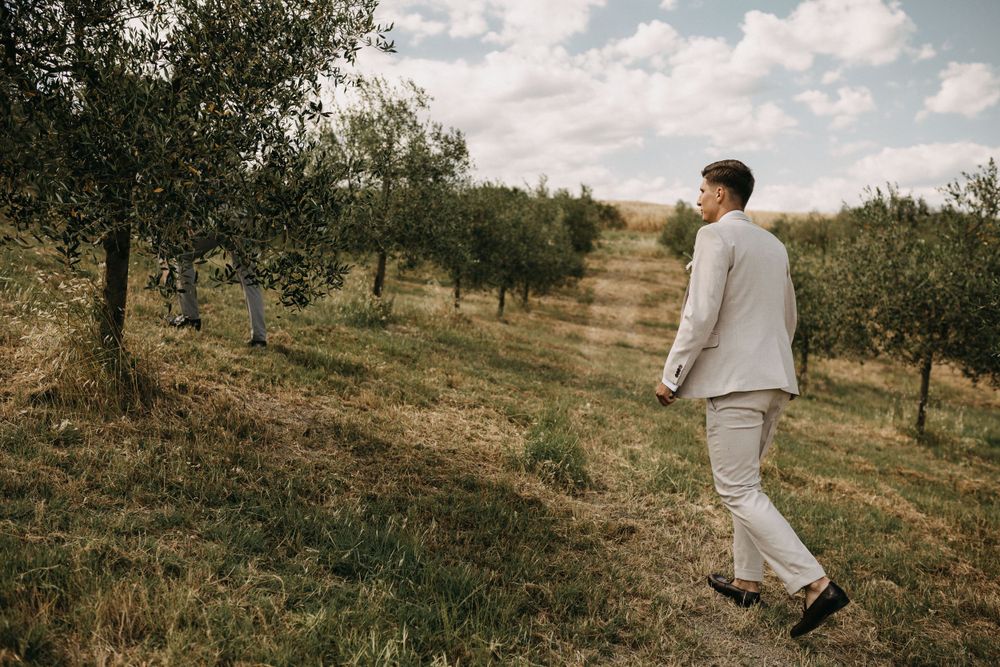 A person in light colored clothing walks through a scenic olive grove on a hillside under cloudy skies.