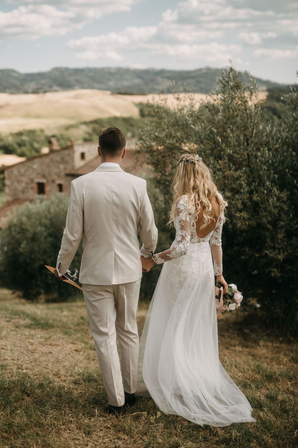 Newlyweds walk hand in hand through an Italian countryside estate surrounded by olive trees.