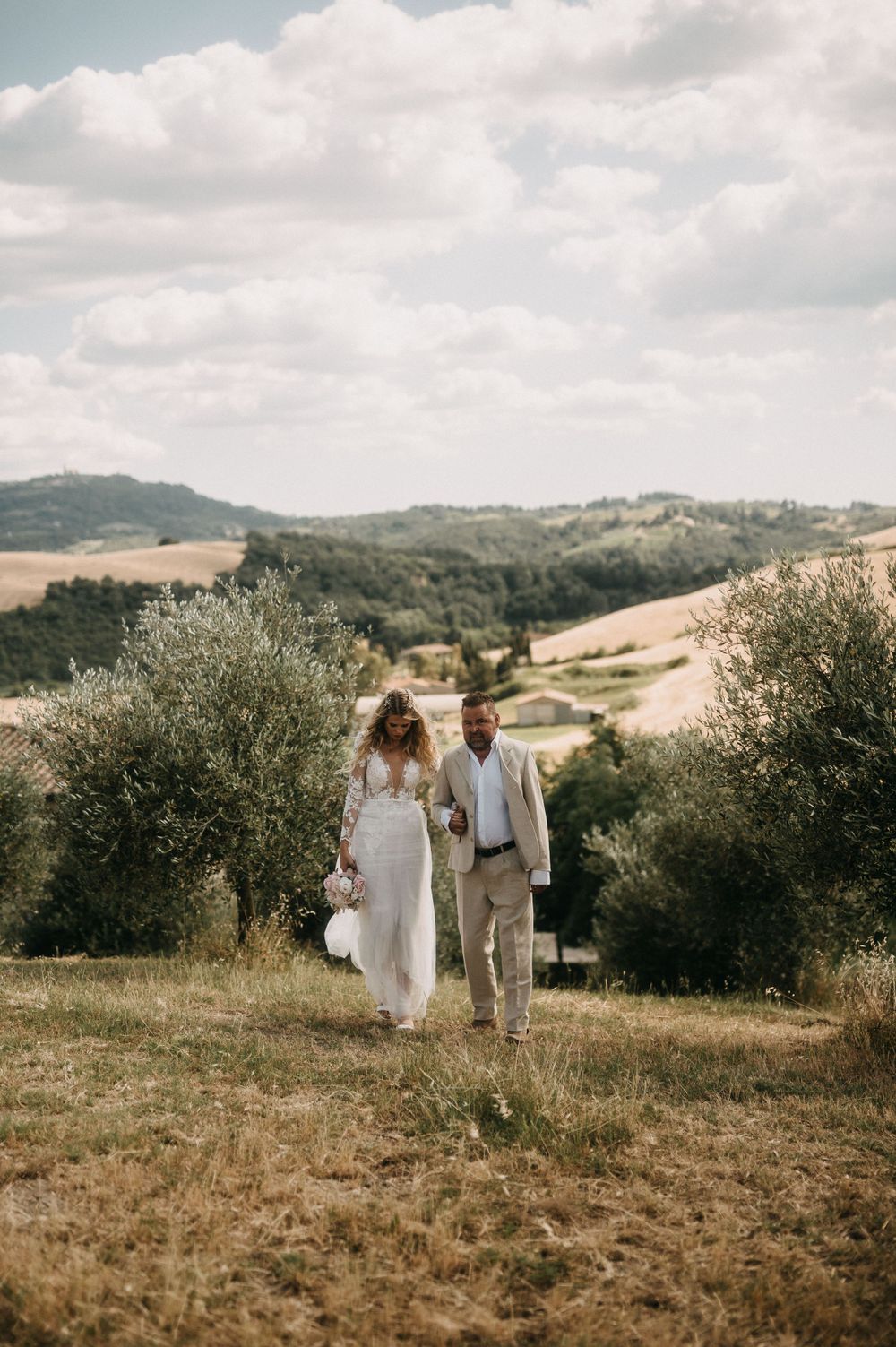 A couple in wedding attire walks through a rustic olive grove landscape in the Italian countryside.