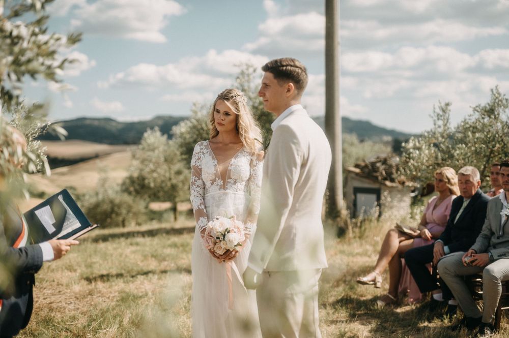 An intimate wedding ceremony takes place in an olive grove with guests seated outdoors.