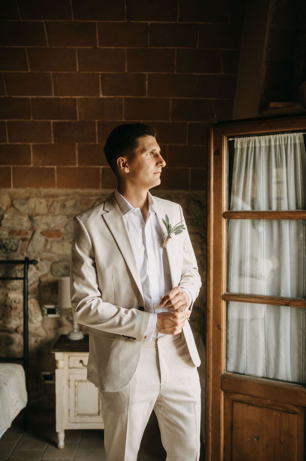 A groom in a beige suit gazes thoughtfully through a window in a rustic stone building.