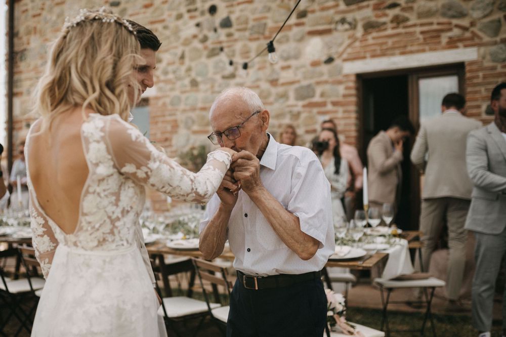 Bride in lace dress sharing an emotional dance moment with elderly gentleman at rustic wedding reception.