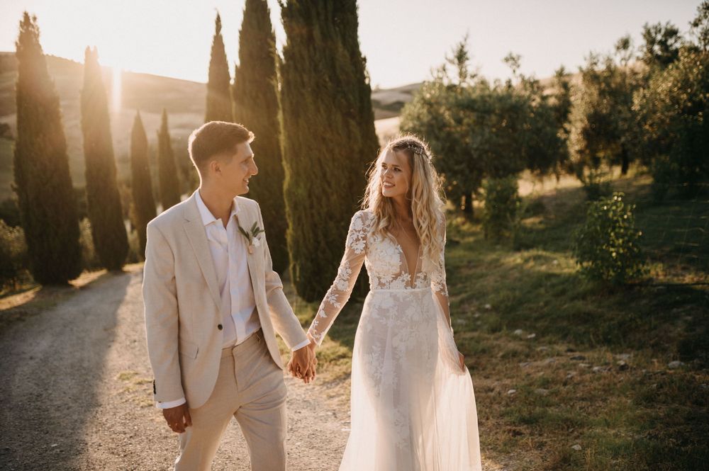 Bride and groom in light-colored attire walking hand in hand along cypress tree path at sunset.