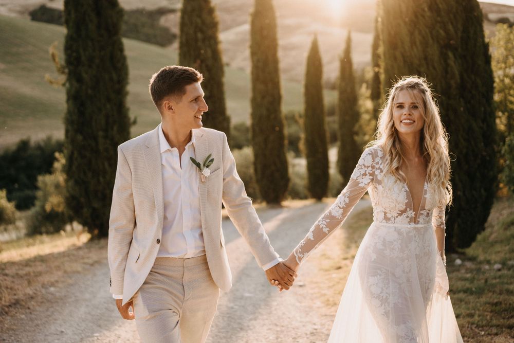Newlyweds strolling together on rustic path lined with Italian cypress trees.