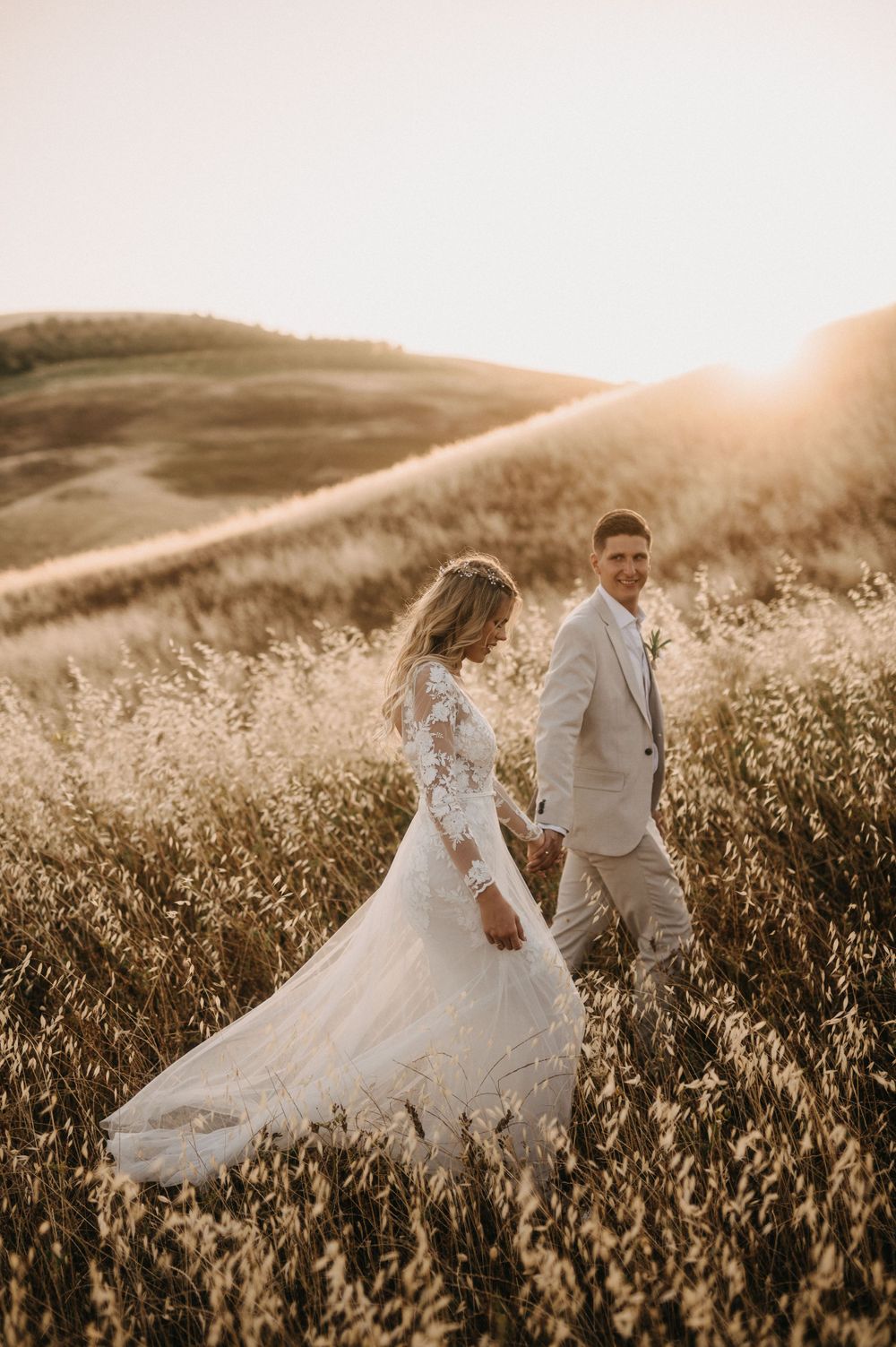 Wedding couple walking through sunlit meadow with flowing bridal gown and light suit.