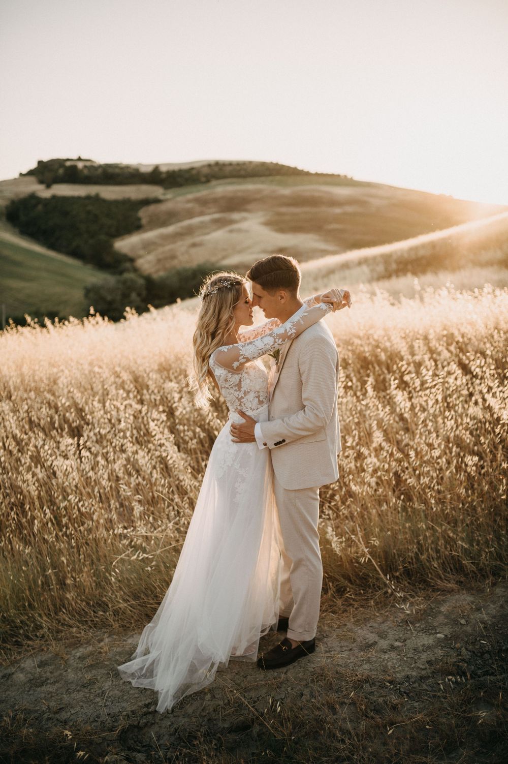Newlyweds embrace in golden sunset light among rolling hills and tall grass.