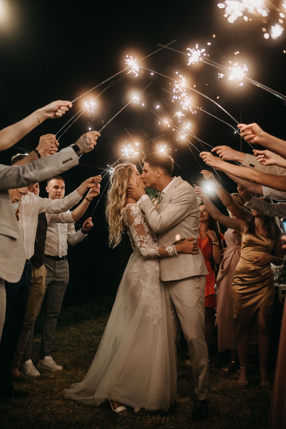 Newlyweds share a romantic moment under sparklers held by wedding guests at night.