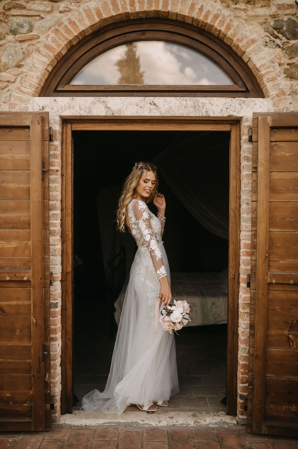 A bride in a lace wedding gown poses in a rustic stone doorway holding a bouquet.