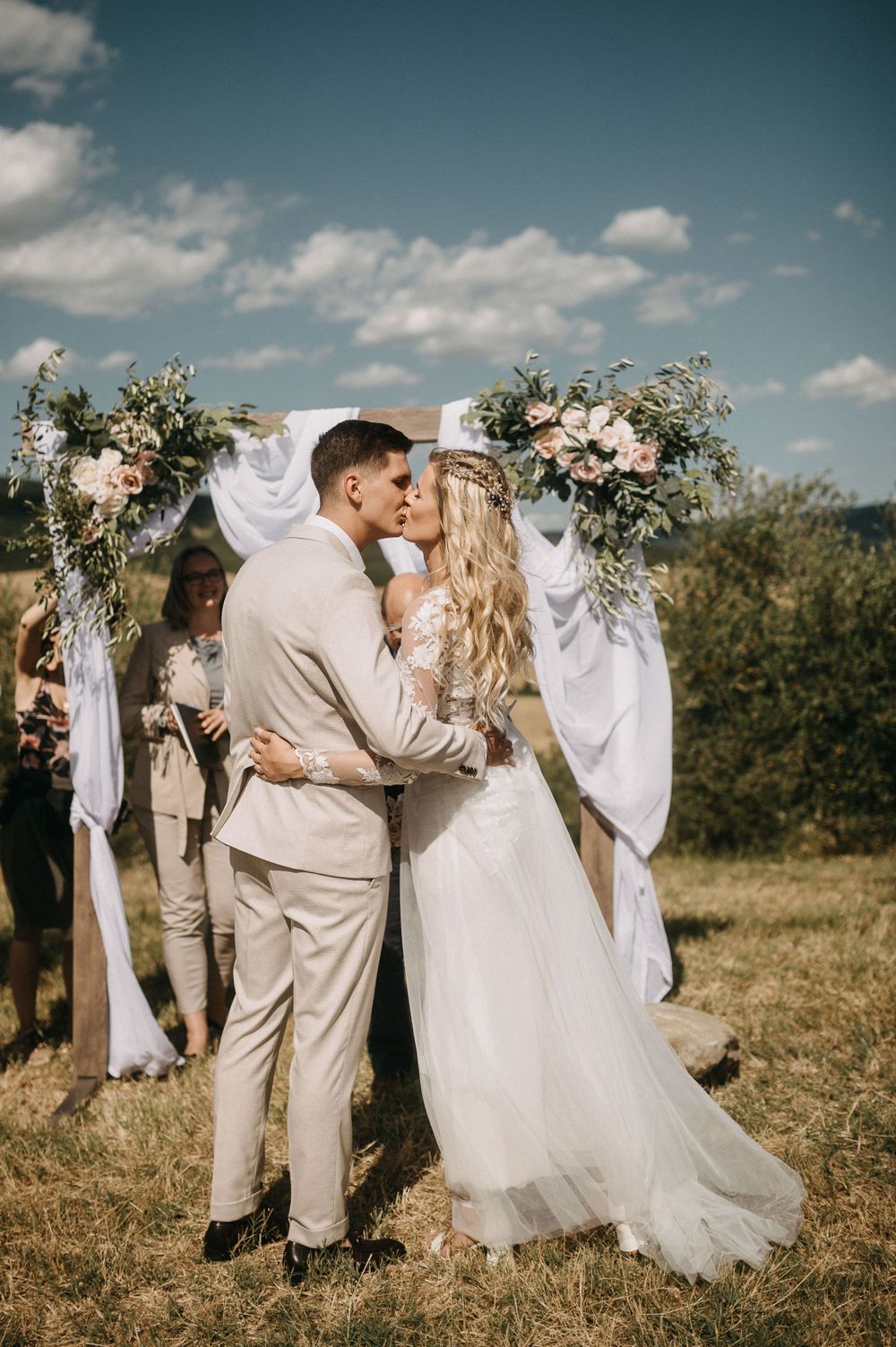 A couple shares their first kiss under a floral wedding arch in a scenic countryside setting.