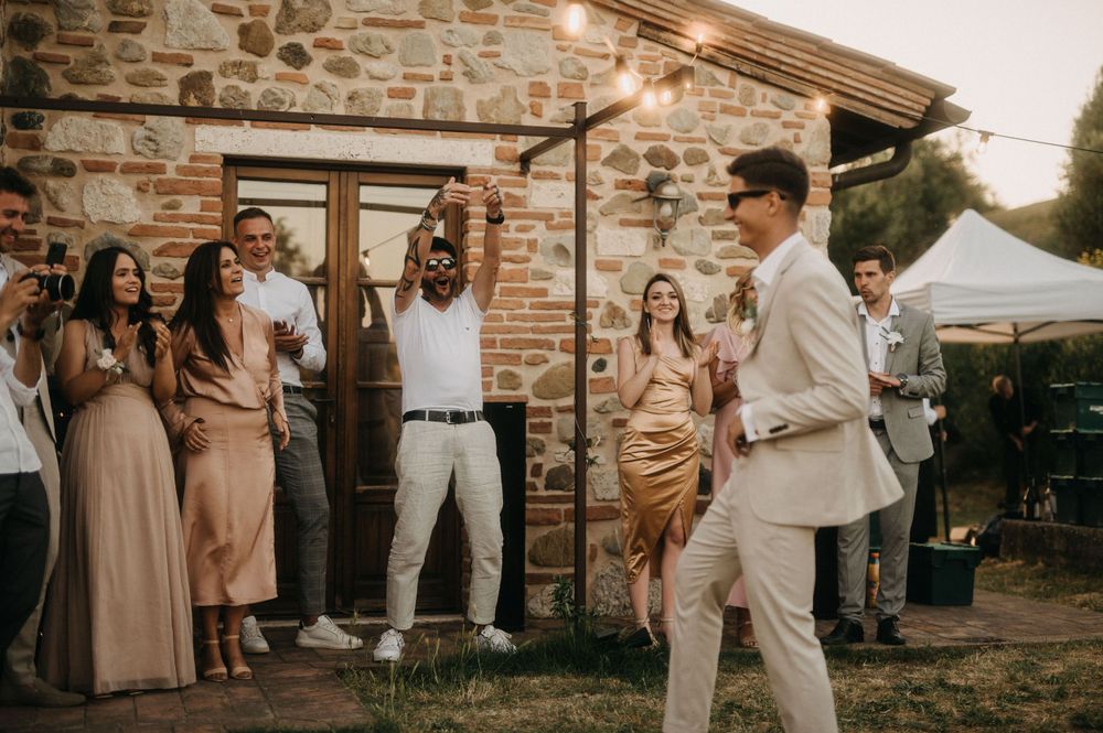 Wedding party celebrates with dancing and festivities outside rustic stone villa with string lights at sunset.