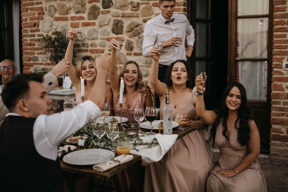 Wedding reception guests toast and celebrate at dinner table against brick wall backdrop.