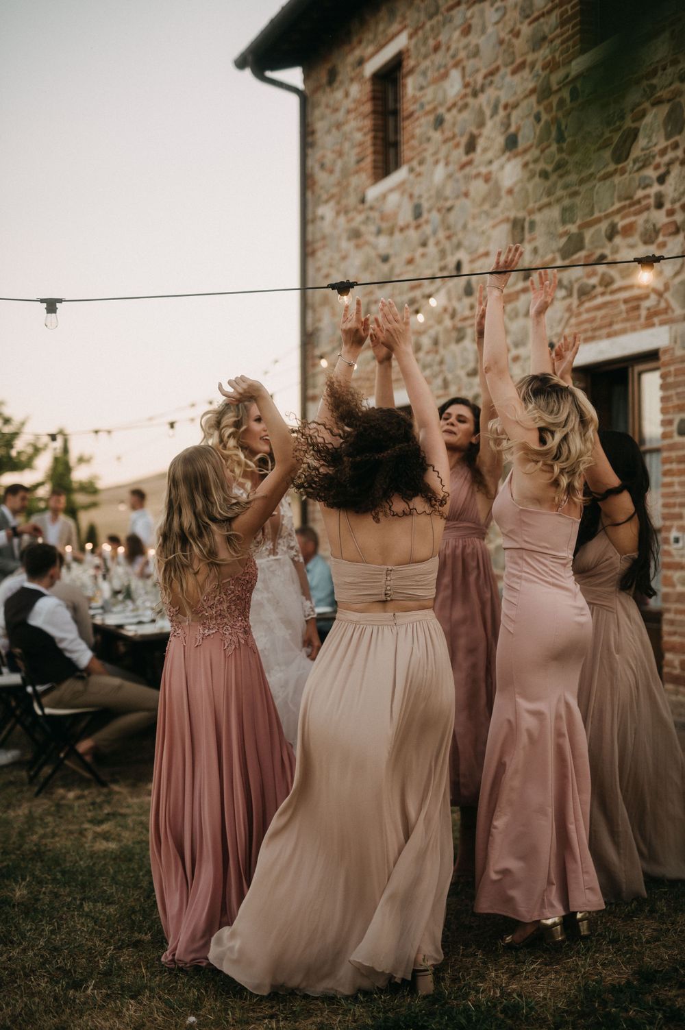 Group wearing blush and neutral gowns dances together under string lights by stone building.