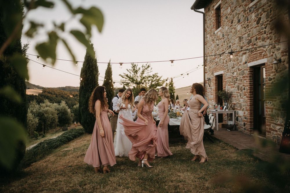 Group in flowing blush dresses dance together on lawn by rustic stone building.