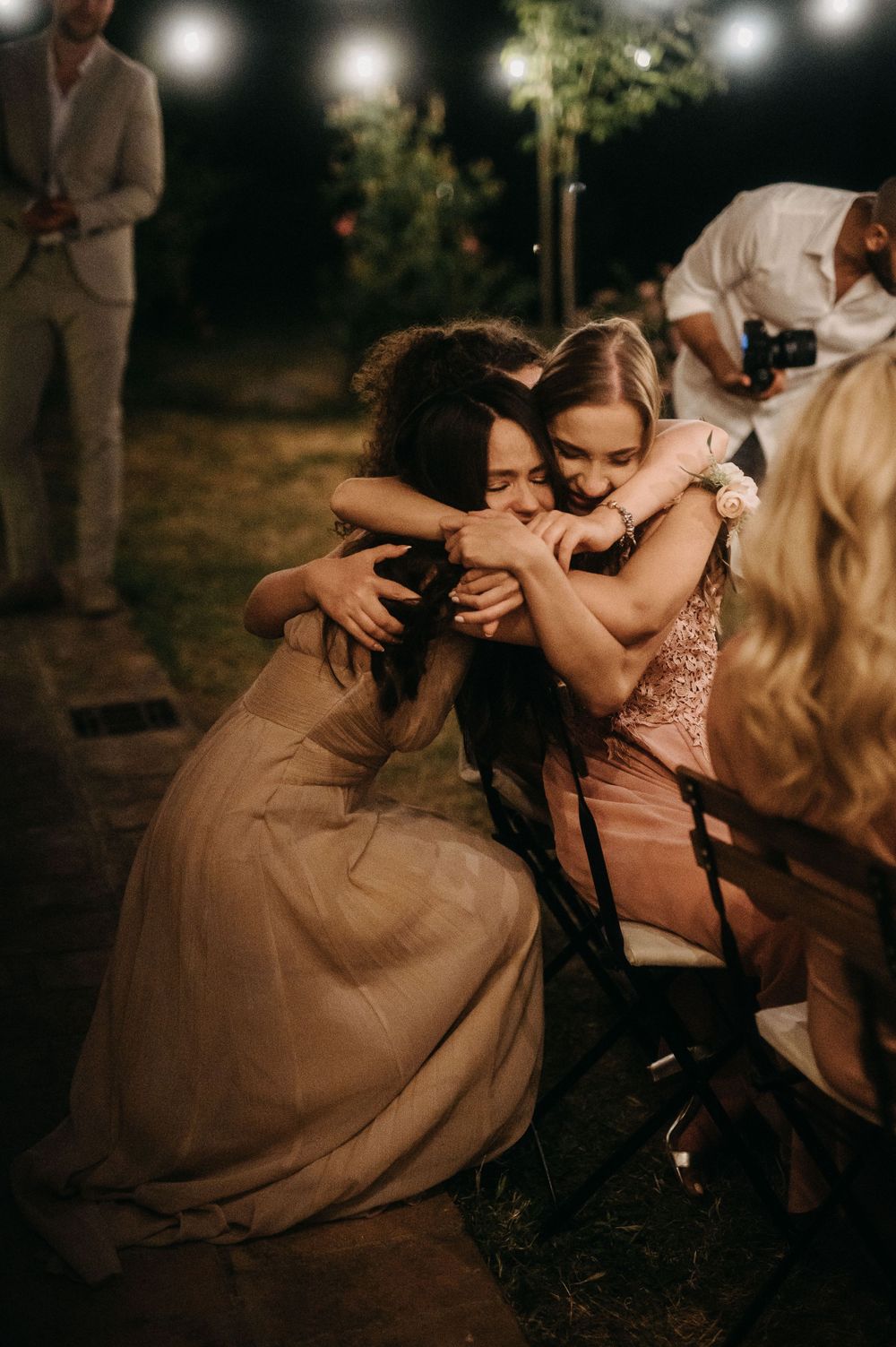 Friends sharing emotional moment at evening wedding reception under string lights.
