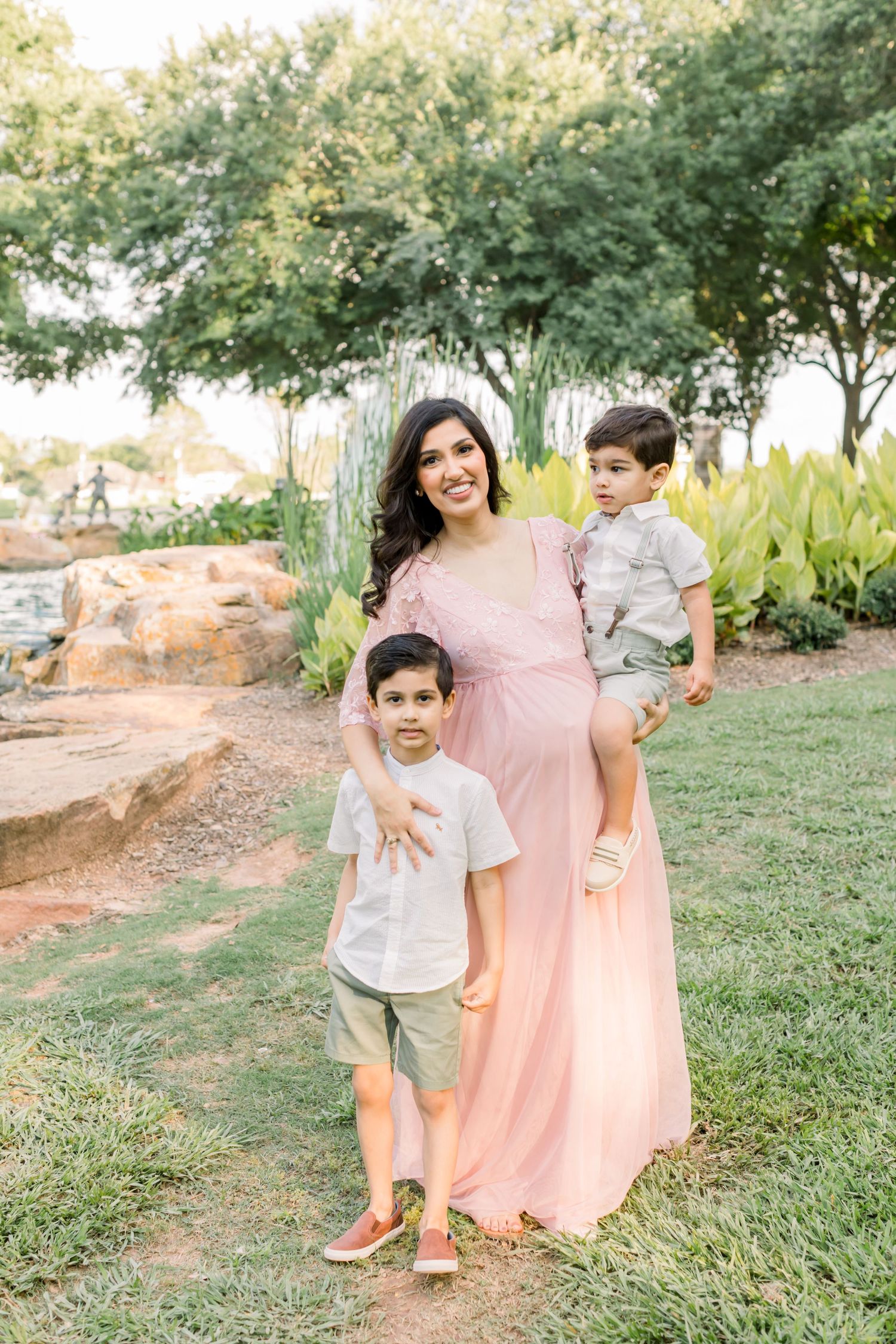 A woman in a pink dress stands with two children on grass with trees and rocks in the background.