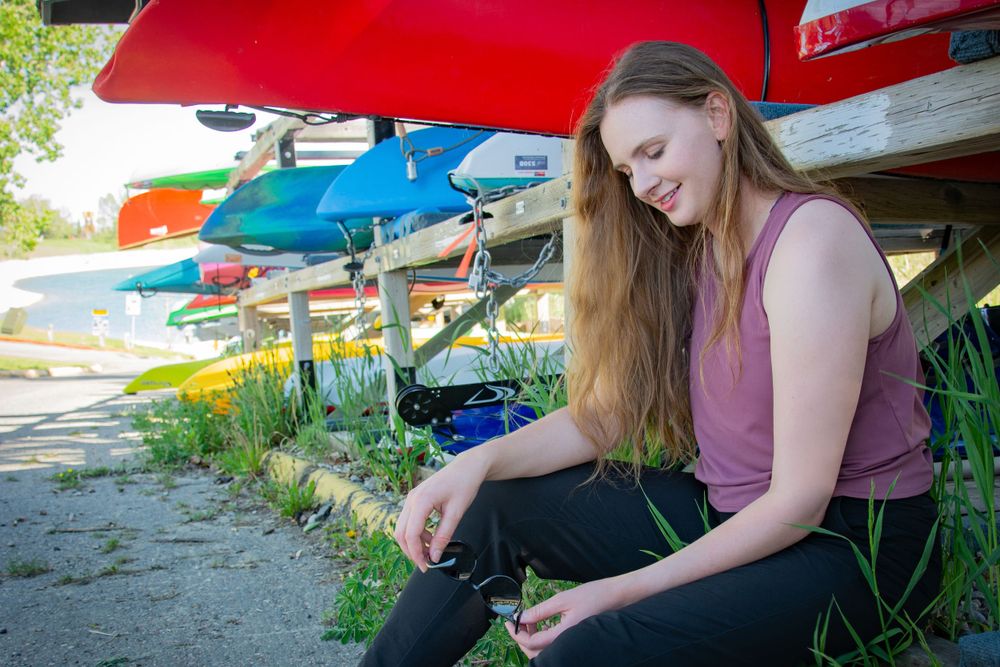 woman poses next to canoe in summer while holding sunglasses smiling and looking down in calgary